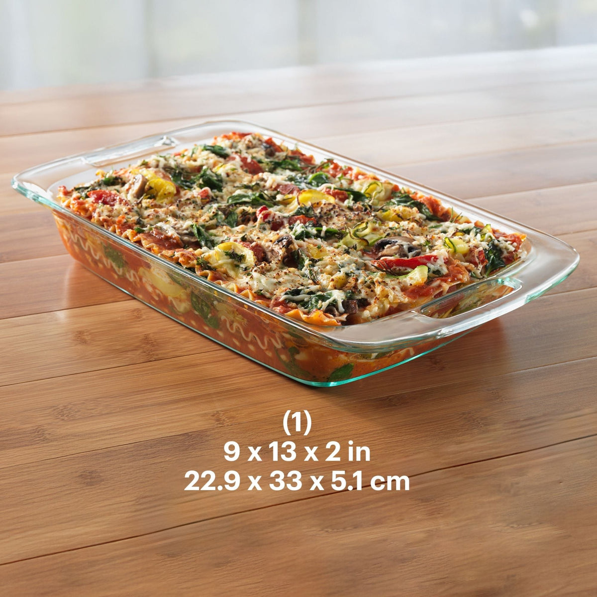 Glass baking dish with a lasagna on a wooden table, showing dimensions.