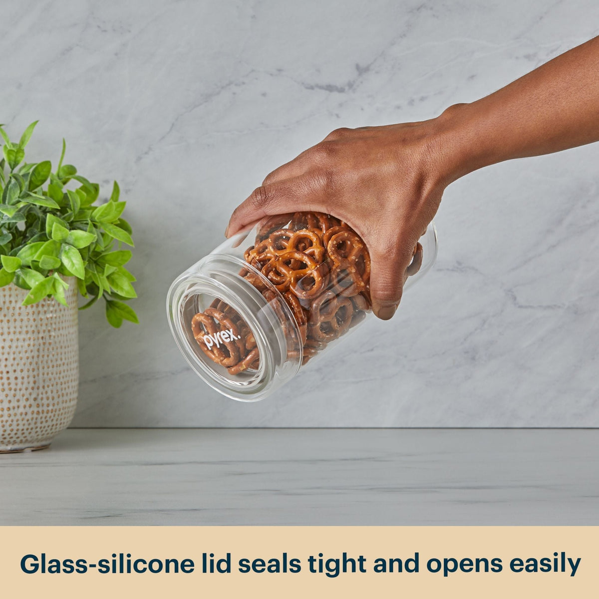 Hand holding a Pyrex jar with pretzels next to a plant on a light gray surface.