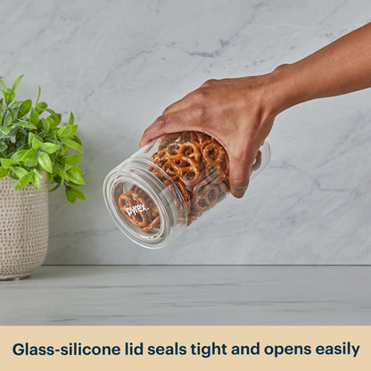 Hand holding a Pyrex jar with pretzels next to a plant on a light gray surface.